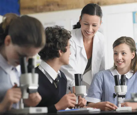 Students look through microscopes while a teacher observes and assists in a classroom setting.