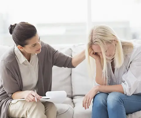A therapist comforts a distressed woman sitting on a couch in a bright, airy room.