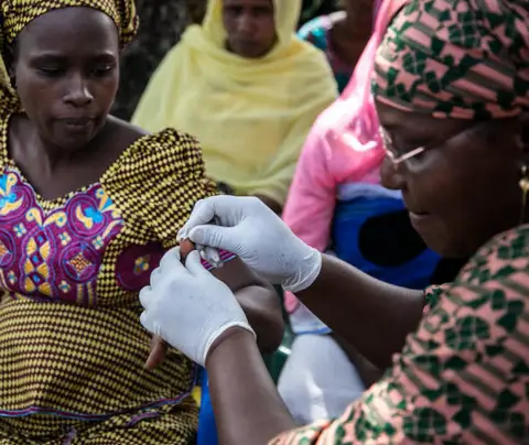 A woman in gloves administers a health treatment to another, surrounded by attentive onlookers.