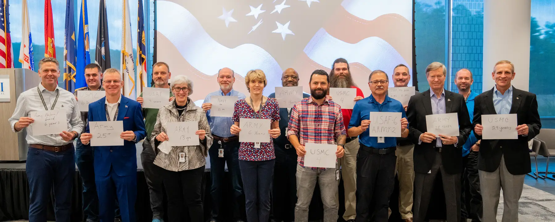 A group of individuals holds signs displaying military service years, surrounded by flags.