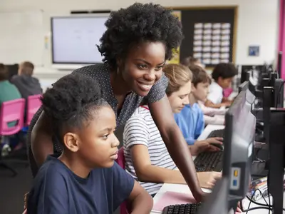 Teacher Helping Female Pupil Line Of High School Students Working at Screens In Computer Class