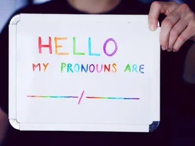 Photo of someone holding a whiteboard containing the words "Hello, my pronouns are" in rainbow colors