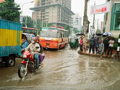Flooding in Dhaka. People standing on the sidewalk. A motorcycle riding through the flooding.