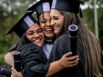 Three graduates in caps and gowns hugging and smiling while holding their diplomas.