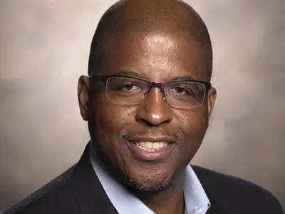 A smiling man with glasses poses in a professional setting, wearing a blazer over a shirt.