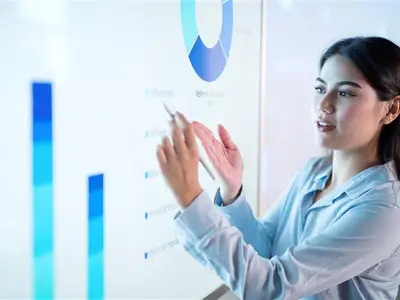 Photo of a young woman pointing at a large screen with graphs and charts