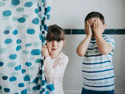 Two kids in a bathtub fully clothed and appearing around the corner of the shower curtain