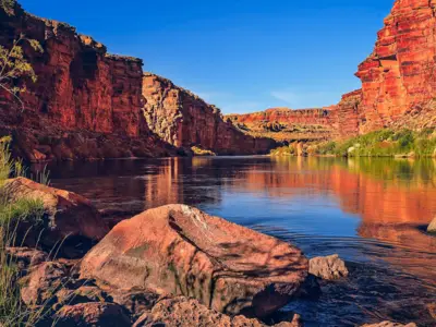 River flowing between steep, red canyon walls with greenery and rocks along the water's edge.
