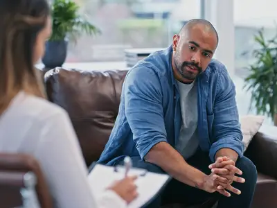 Man sitting on a couch talking to a woman with a notepad in an office setting.