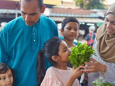 Family buying fresh vegetables at a market, surrounded by stalls and other shoppers.