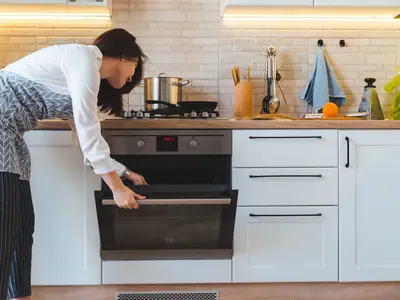 Woman opening oven door in modern kitchen with utensils and appliances on the countertop.