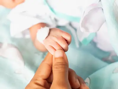 Baby's bandaged hand grasping an adult finger, lying on a light blue blanket.