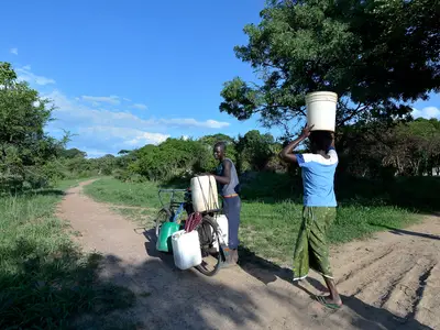 Two people transporting water containers, one on a bicycle, another balancing one on her head, along a dirt path surrounded by greenery.