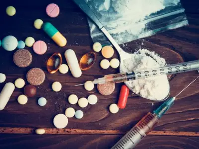 Various pills and syringes lie scattered on a wooden surface next to a bag of white powder.