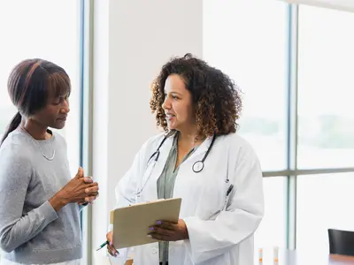 Doctor holding a clipboard, conversing with a patient in a bright, windowed medical office.
