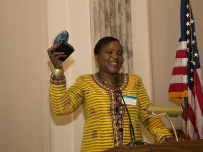 Smiling woman holding an award behind a podium beside an American flag in a formal setting.