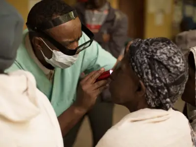 Healthcare worker checks patient's eyes with flashlight in a crowded indoor clinic.