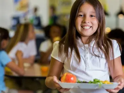 A girl smiles and holds a lunch tray in a school cafeteria with other children.