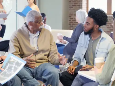 People having a discussion while holding documents in a spacious, well-lit room with other groups.