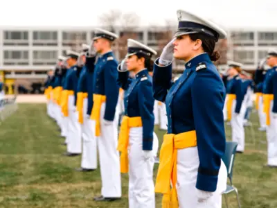 Cadets in blue uniforms and yellow sashes salute, standing in rows on a grassy field.