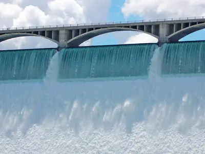 Water cascades over a dam, above which an arched bridge spans against a cloudy sky backdrop.
