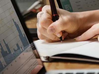 Woman's hand is seen writing in a notebook while computer screens displaying different data are on either side.