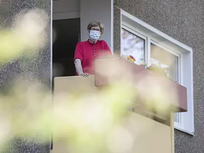 A senior woman wearing a mask looks out from a balcony.
