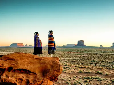 Two Navajo sisters in traditional clothing watch a sunset in Monument Valley.