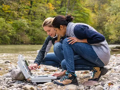 Two female scientists look at data on a tablet next to a creek.
