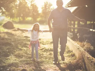 A smiling granddaughter and grandfather walk near a barn on a farm.