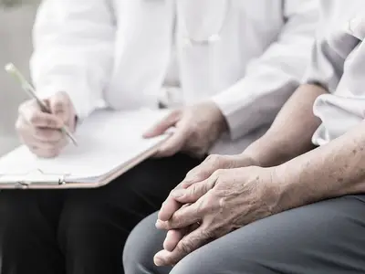 A medical professional in a white coat takes notes on a clipboard while talking to a senior patient.