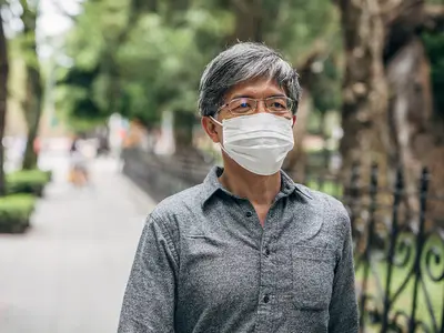 An Asian senior man wears a face mask and walks along a tree-lined sidewalk.