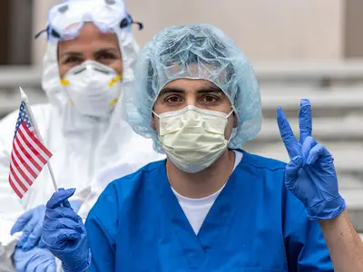 A male health worker in protective gear holds an American flag and flashes the "V for Victory" hand sign.