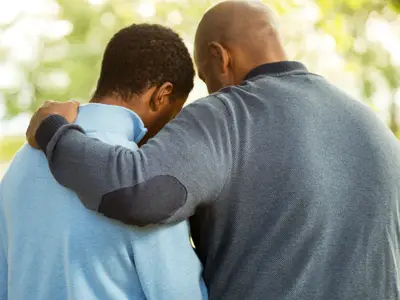 An African-American father places his arm around the shoulders of his adult son.