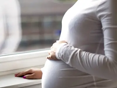 Pregnant woman stands holding belly, gazing out large window at blurred cityscape.
