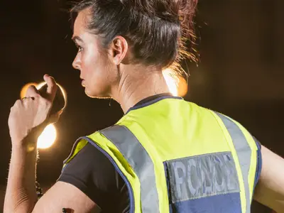 A female police officer talks into a handheld radio while watching traffic on a street at night.