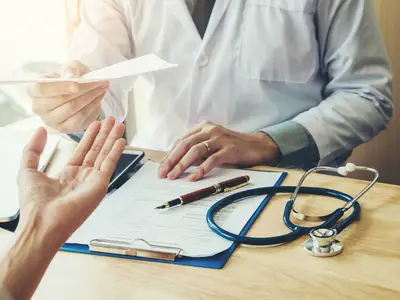 A doctor hands a prescription form to a patient across a desk.