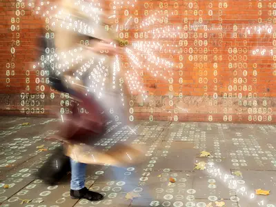 A blurred image of a female shopper walking along a sidewalk with a phone, with digits superimposed on the image to suggest data traveling to and from the phone.