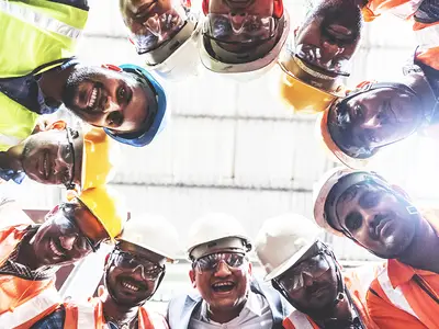 A group of engineers in hard hats at an Indian plant form a circle and smile for the camera.