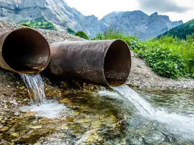 Two large pipes releasing water into a stream in a mountainous, grassy landscape.