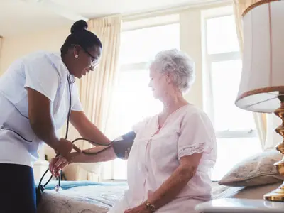 Nurse taking elderly woman's blood pressure in a sunlit bedroom with bed and lamp.