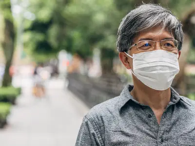 An Asian senior man wears a face mask and walks along a tree-lined sidewalk.