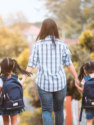 Photo of a mother holding hands with her two children wearing backpacks, walking away from the camera