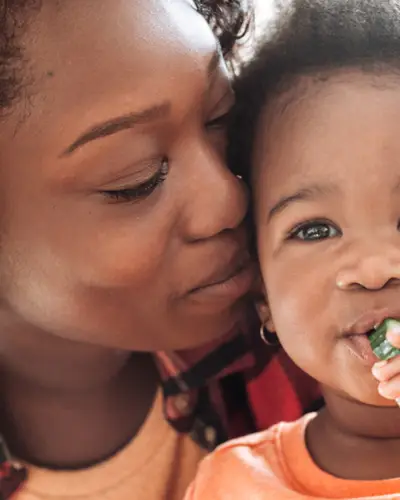 A mother holds her toddler, who is eating a slice of cucumber.