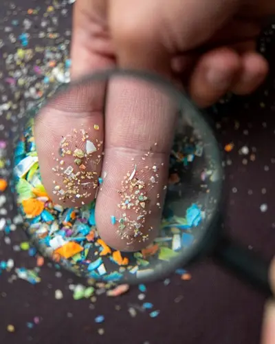 Small chips of plastic waste are scattered on a flat surface. Another small amount rests on the tips of two fingers and is enlarged by a round magnifying glass.