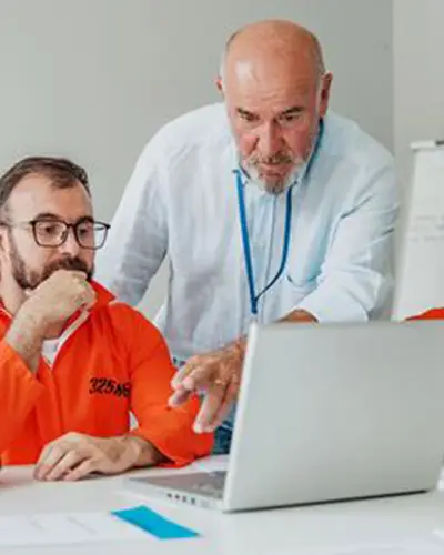 Incarcerated men looking at a laptop. An instructor pointing to the laptop's screen