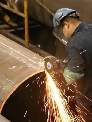 Worker using an angle grinder on a large metal pipe, surrounded by industrial equipment and pipes.