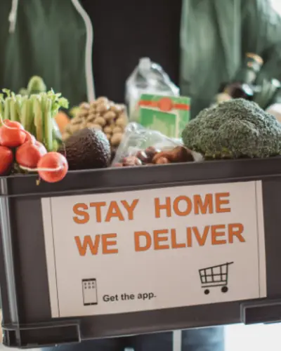 Person holding a crate of groceries with "STAY HOME WE DELIVER" printed on it.
