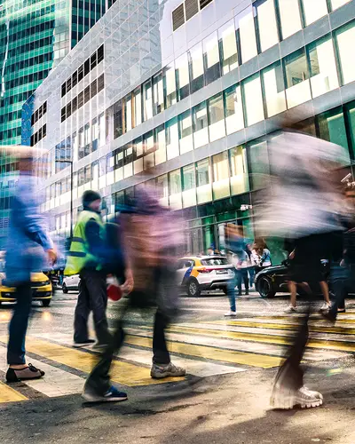 An evening city scene with pedestrians crossing a busy intersection.