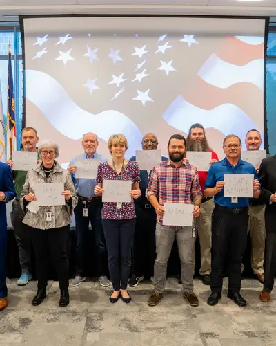 A group of individuals holds signs displaying military service years, surrounded by flags.